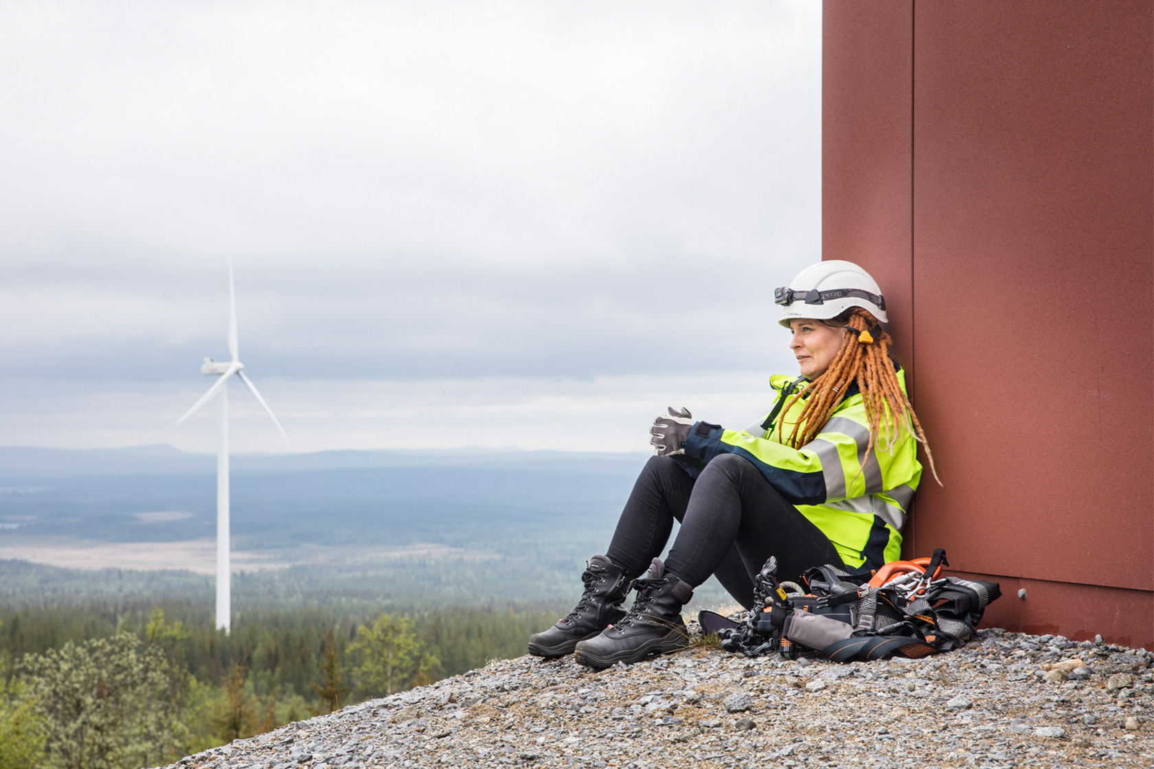 En kvinna i hjälm som sitter lutad mot en vägg och blickar ut mot ett vindkraftverk