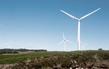 Image of a wind turbine located on a hill, in a green landscape with blue skies above.