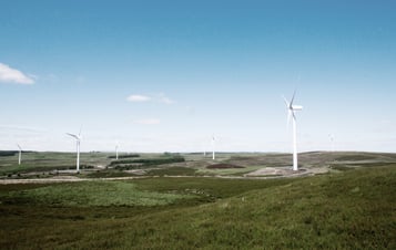 Photo of wind turbines located in a green landscape, with blue skies above.