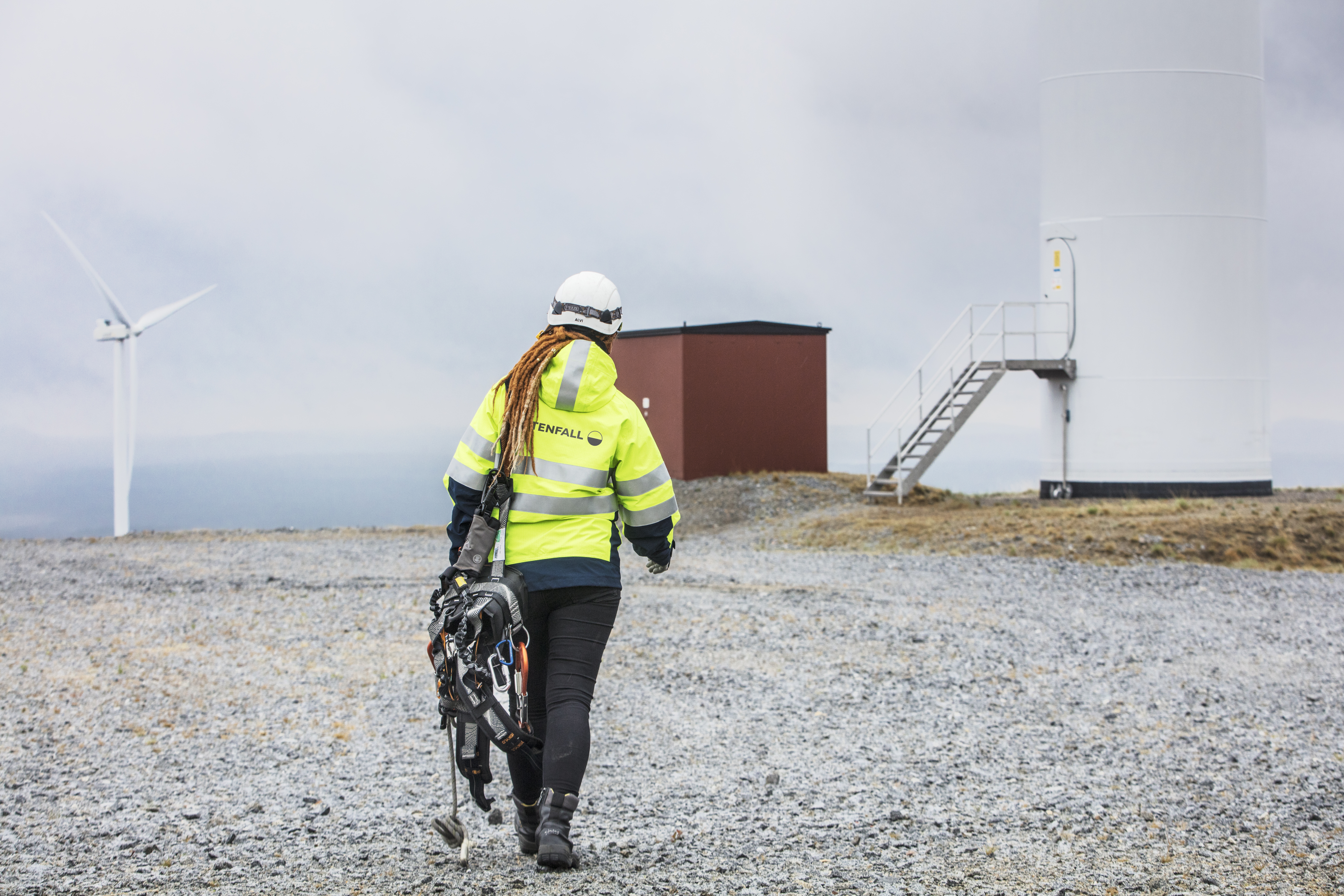Image of a Vattenfall employee wearing safety clothes and helmet, walking across dry ground towards a wind turbine.