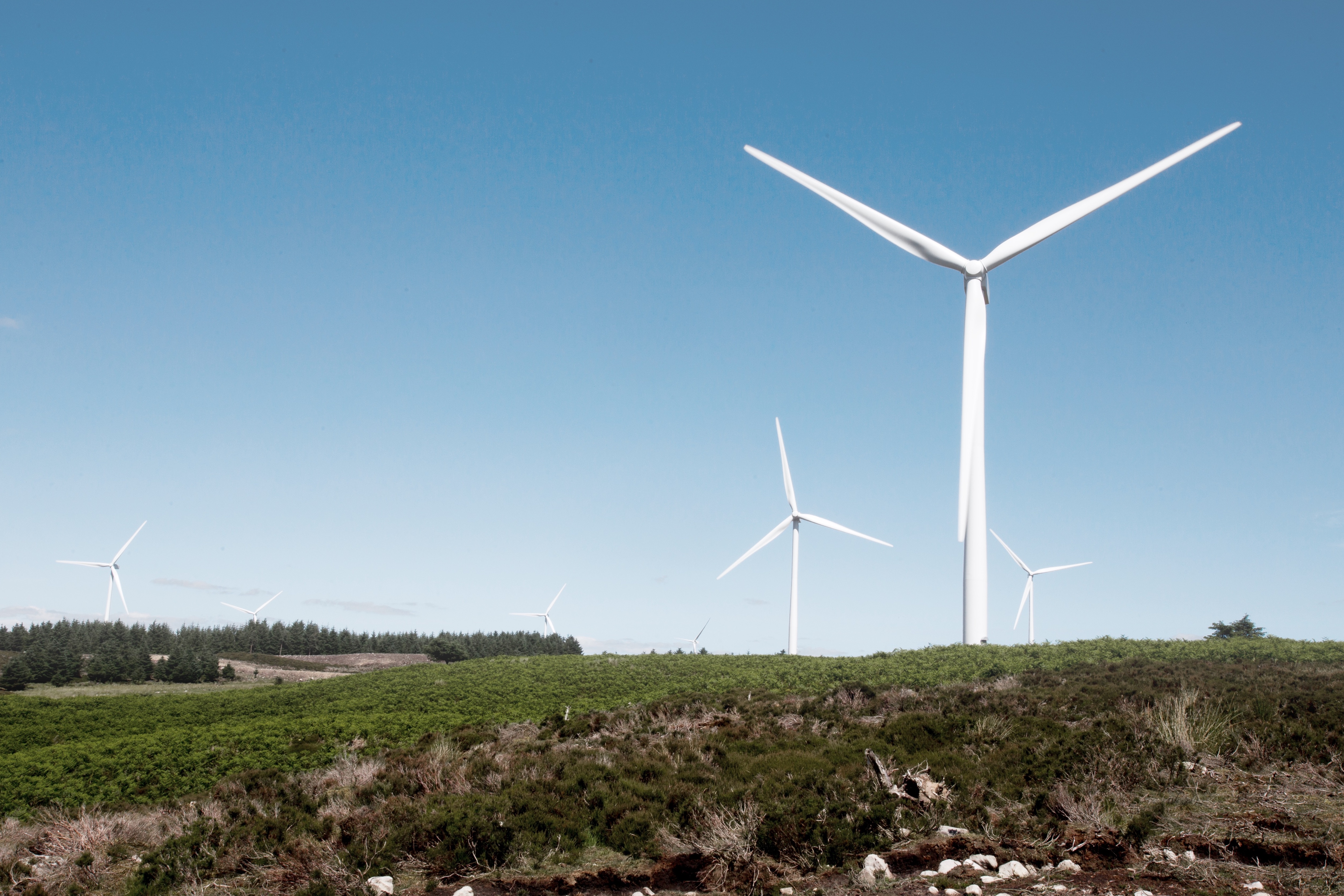 Image of a wind turbine located on a hill, in a green landscape with blue skies above.