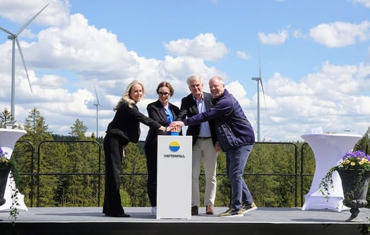 Representatives from Vattenfall, Eksjö Municipality and Volvo press a button on a podium bearing the Vattenfall logo, on an outdoor stage with wind turbines and a forest in the background. Representatives from Vattenfall, Eksjö Municipality and Volvo press a button on a podium bearing the Vattenfall logo, on an outdoor stage with wind turbines and a forest in the background.