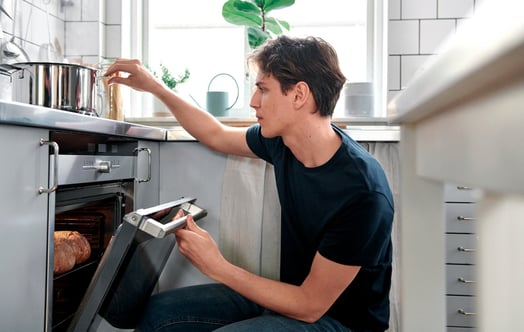 A man kneeling in a bright, modern kitchen, checking food in the oven. A man kneeling in a bright, modern kitchen, checking food in the oven.