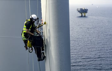 Mann in Sicherheitsausrüstung klettert auf einen Windradflügel einer Offshore-Windkraftanlage Mann in Sicherheitsausrüstung klettert auf einen Windradflügel einer Offshore-Windkraftanlage