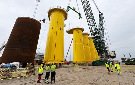 Large offshore wind transition pieces standing in a port area, with cranes and workers nearby. Large offshore wind transition pieces standing in a port area, with cranes and workers nearby.