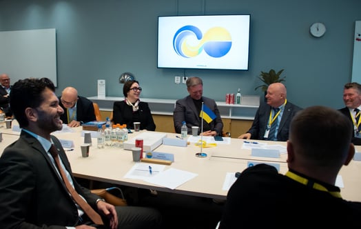 Deltagare sitter runt ett konferensbord under ett möte, med en liten ukrainsk flagga på bordet och en blågul grafik på en skärm. Deltagare sitter runt ett konferensbord under ett möte, med en liten ukrainsk flagga på bordet och en blågul grafik på en skärm.