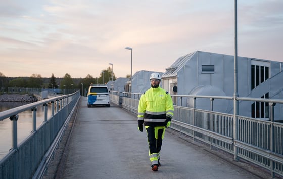 En man i varselkläder och hjälm går på bro intill vattenkraftverk. En man i varselkläder och hjälm går på bro intill vattenkraftverk.